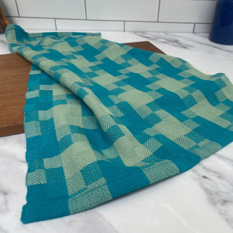 Product photo of teal and pale green checkered towel on a marble surface with a white tiled wall background